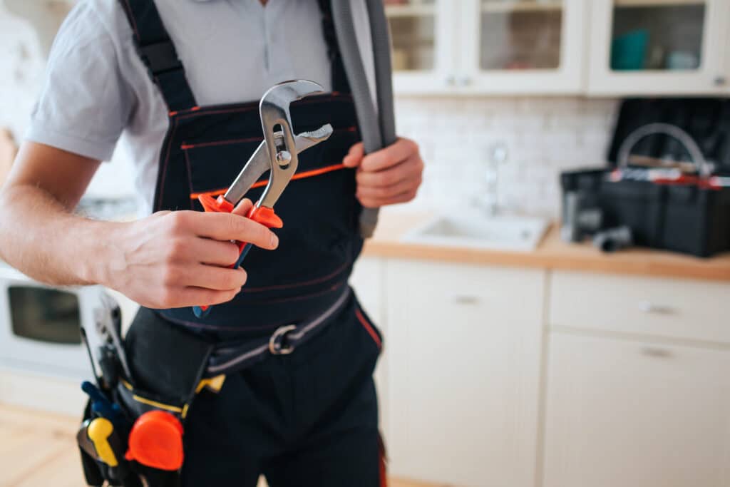 Plumbing service in Conway, SC, a man wearing a working uniform, holds a wrench as he stands in a kitchen during daylight. He has tools on his belt and a toolbox on the desk.